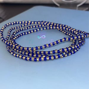May include: A blue and gold beaded waist bead, also known as a waist chain, is made of small, round beads. The beads are arranged in a pattern of alternating blue and gold beads.