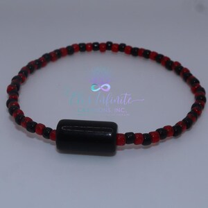 May include: A beaded bracelet featuring alternating red and black beads. A larger, cylindrical black bead is centered on the bracelet. The bracelet is likely handmade and is displayed on a light background. The text "The Infinite Creations, Inc." is visible.