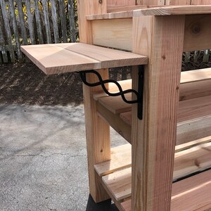 Puede incluir: Una mesa de trabajo de madera con un estante y un estante peque&ntilde;o y estrecho unido al lado. El estante peque&ntilde;o est&aacute; sostenido por un soporte de metal negro.