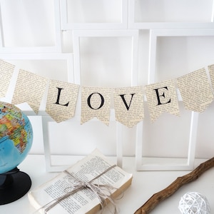 May include: A banner made from book pages with the word "LOVE" spelled out in black letters. The banner is hanging between white picture frames and a globe is in the foreground.