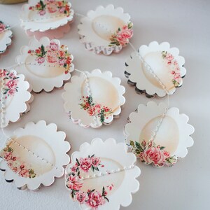 May include: A garland of scalloped paper cutouts, each adorned with a floral design featuring pink roses and greenery. The cutouts are connected by a white string, creating a decorative banner. The background is white.