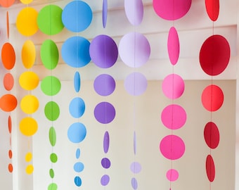 Könnte beinhalten: Eine farbenfrohe Papiergirlande mit kreisförmigen Ausschnitten in Regenbogenfarben. Die Girlande zeigt ein Spektrum an Farben, darunter Rot, Orange, Gelb, Grün, Blau, Lila und Rosa. Die Kreise sind an einer Schnur aufgehängt und bilden eine dekorative Hängedekoration.