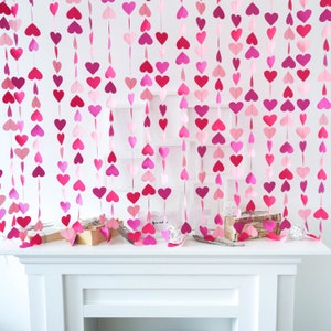 Può includere: Ghirlanda di cuori di carta rosa e rossa appesa a un muro bianco. La ghirlanda è composta da più fili di cuori, ogni filo è di una tonalità diversa di rosa o rosso.