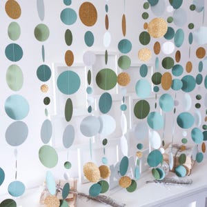 May include: Decorative paper garland featuring a variety of circular and oval shapes in shades of teal, green, gold glitter, and white. The garland is hanging in front of a white wall with framed artwork.