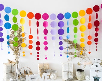 Könnte beinhalten: Eine farbenfrohe Regenbogen-Girlande aus Papierkreisen hängt vor einer weißen Wand. Die Girlande besteht aus roten, orangen, gelben, grünen, blauen und lila Kreisen. Dekorative Gegenstände sind auf einer weißen Oberfläche darunter angeordnet.