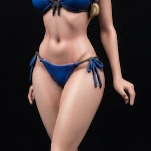 Può includere: Una statuetta in piedi di un personaggio biondo con un'acconciatura a treccia e una fascia scura. La figura indossa un bikini blu a due pezzi con dettagli a laccetti sui fianchi dello slip e un top semplice. Il personaggio ha occhi azzurri.