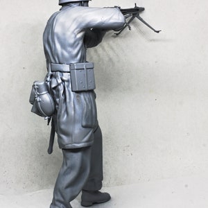 May include: A gray plastic model of a soldier in a helmet holding a machine gun. The soldier is wearing a uniform and has a belt with a pouch attached. The soldier is standing on a gray background.