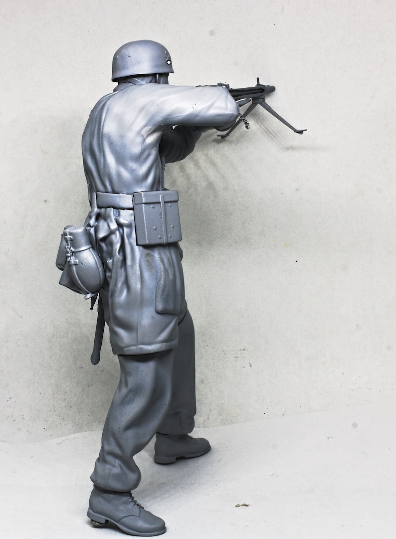 May include: A gray plastic model of a soldier in a helmet holding a machine gun. The soldier is wearing a uniform and has a belt with a pouch attached. The soldier is standing on a gray background.
