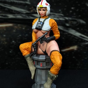 May include: A female action figure wearing an orange and white flight suit with a helmet and holding a blaster. The figure is sitting on a gray platform with red lights.