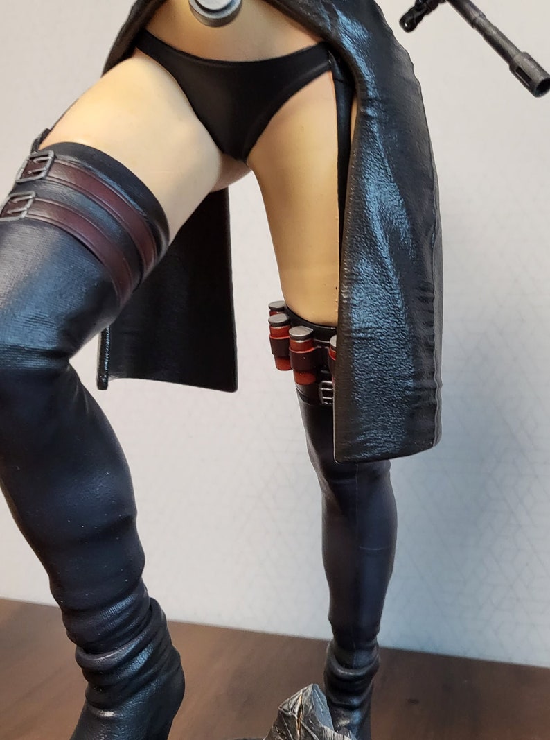 May include: A black leather figure with a black and red belt and a black leather skirt. The figure is wearing black leather boots and has a black leather strap on the left leg.