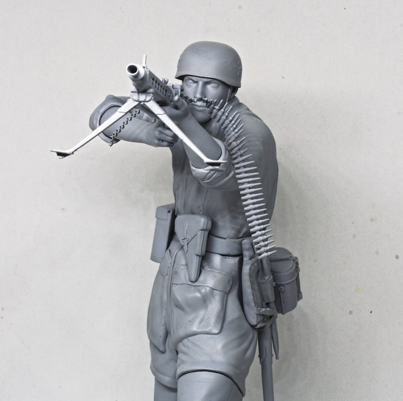 May include: A gray plastic model of a soldier holding a machine gun. The soldier is wearing a helmet and has a belt of ammunition around his waist.