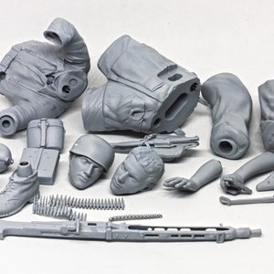 May include: A gray plastic model kit of a soldier, featuring a torso, legs, arms, head, and accessories like a machine gun, ammunition belt, and a helmet. The kit is unassembled and ready for painting.