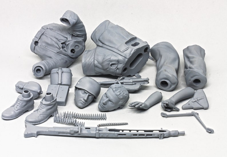 May include: A gray plastic model kit of a soldier, featuring a torso, legs, arms, head, and accessories like a machine gun, ammunition belt, and a helmet. The kit is unassembled and ready for painting.