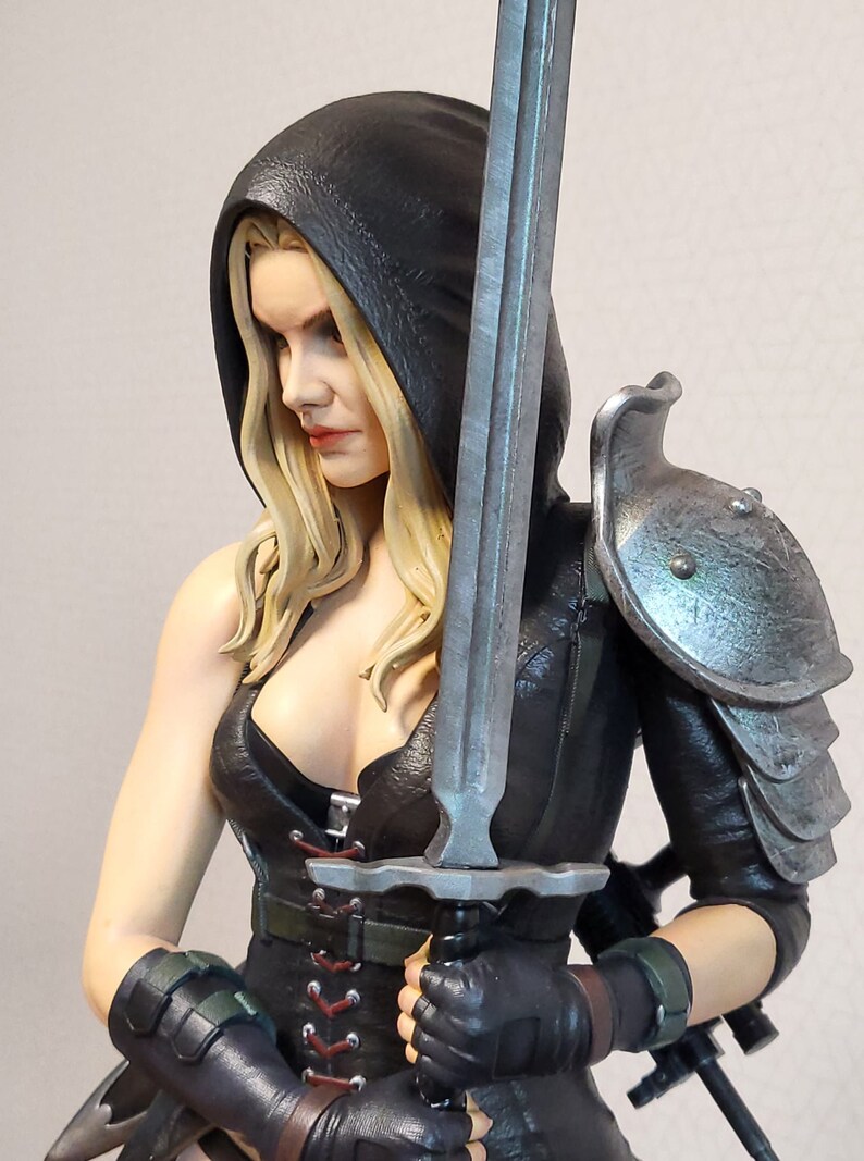 May include: A female action figure with long blonde hair, wearing a black leather outfit with a hood and holding a silver sword. She is wearing silver metal shoulder armour.