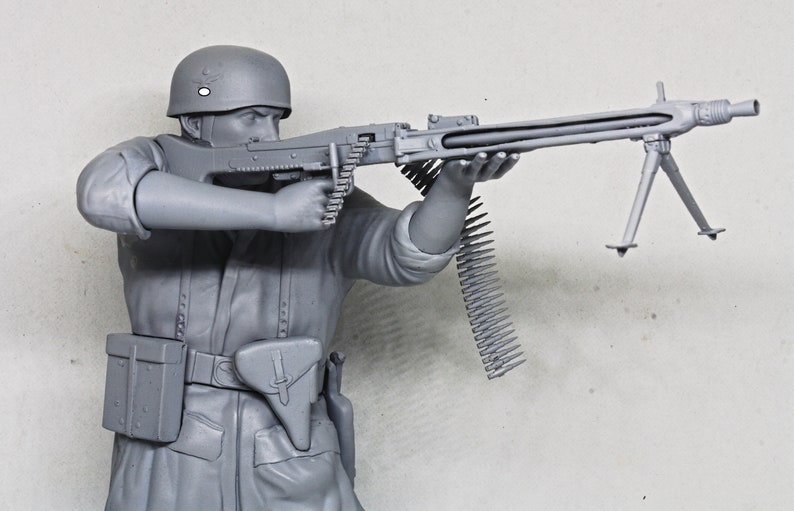 May include: A gray plastic model of a soldier holding a machine gun with a bipod. The soldier is wearing a helmet and a belt with a pouch. The machine gun has a long barrel and a belt of ammunition.