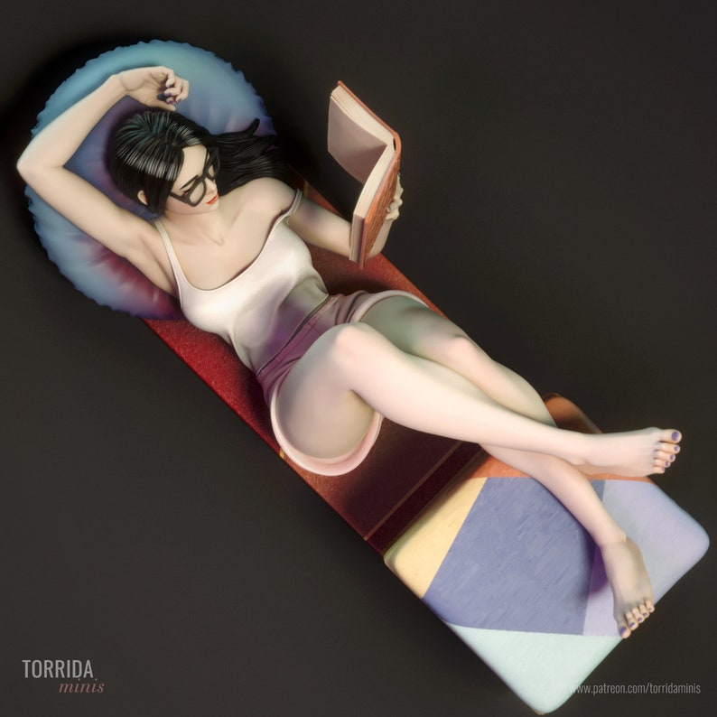 May include: A woman in a white vest top and a purple skirt is lying on a colourful patterned cushion, reading a book. She has long black hair and is wearing glasses.