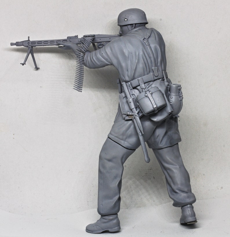 May include: A gray plastic model of a soldier holding a machine gun. The soldier is wearing a helmet and a uniform with a belt and pouches. The machine gun has a long barrel and a bipod.
