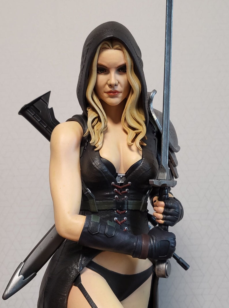 May include: A female action figure with long blonde hair, wearing a black leather outfit with a hooded cloak, holding a silver sword in her right hand. She is wearing black leather gloves and has a serious expression on her face.