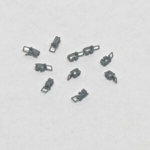 Puede incluir: Diez peque&ntilde;os clips de pl&aacute;stico gris, esparcidos sobre una superficie blanca.