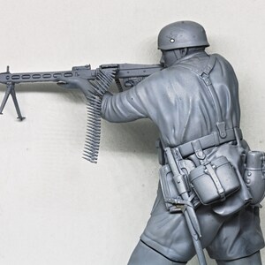 May include: A gray plastic model of a soldier holding a machine gun. The soldier is wearing a helmet and a belt with pouches. The machine gun has a bipod and a long belt of ammunition.