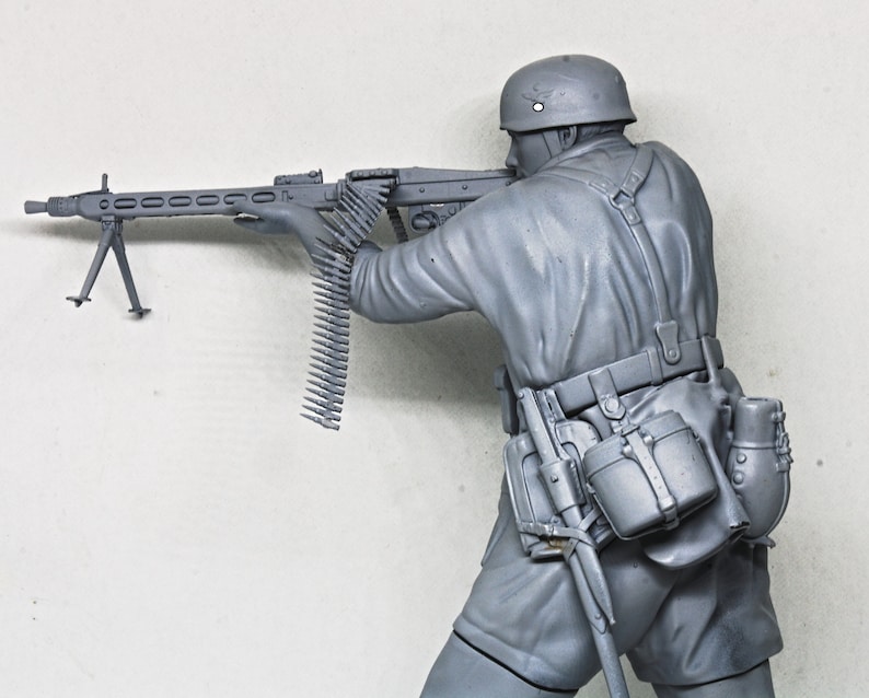 May include: A gray plastic model of a soldier holding a machine gun. The soldier is wearing a helmet and a belt with pouches. The machine gun has a bipod and a long belt of ammunition.