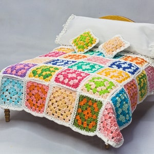May include: A miniature bed with a colourful, crocheted blanket. The blanket is made of squares in various colours, including pink, green, blue, and orange. Two small square pillows and a white pillow with lace trim complete the bed set.