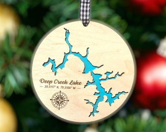 Könnte beinhalten: Ein rundes Holzornament mit einer ausgeschnittenen Karte des Deep Creek Lake, die mit hellblauem Harz gefüllt ist. Der Text lautet Deep Creek Lake 39.5117 N, 79.3156 W, ergänzt durch eine Kompassrosengrafik. Es hängt an einem schwarz-weißen Vichy-Band.