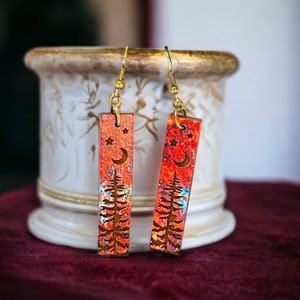 Può includere: Un paio di orecchini in legno con un design rosso, arancione e blu. Gli orecchini presentano una luna crescente e stelle con una silhouette di un pino.