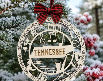 May include: A circular wooden ornament featuring a cutout map of Tennessee with the text TENNESSEE, surrounded by icons of a guitar, drums, music notes, and a grill. The border includes text reading GREAT SMOKY MOUNTAINS, GRACELAND, COUNTRY MUSIC, BARBECUE, HOME OF THE BLUES, and PARTHENON, topped with a red and black plaid ribbon bow.