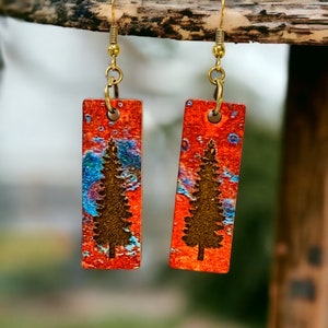 May include: A pair of orange and blue earrings with a silhouette of a pine tree on each. The earrings are made of metal and have a rectangular shape.