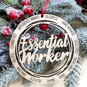 May include: A wooden Christmas ornament with the words "Essential Worker" in the center. The ornament is surrounded by a circle of words, including "Brave", "Selfless", "Amazing", "Helper", "Committed", "Difference Maker", "Sacrifice", "Hard Worker", "Dedicated", "Invaluable", and "Hero".