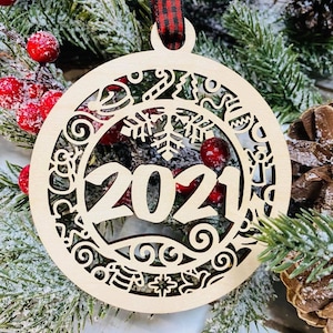 Könnte beinhalten: Ein hölzerner Weihnachtsbaumschmuck mit einem Schneeflockenmuster und dem ausgeschnittenen Jahr 2021. Der Schmuck hängt an einem rot-schwarzen Band.