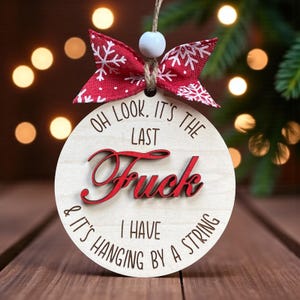 May include: A round wooden Christmas ornament with the text "OH LOOK, IT'S THE LAST FUCK I HAVE & IT'S HANGING BY A STRING" in black and red. It has a red bow with white snowflakes and a string for hanging.