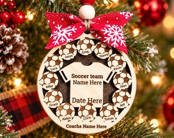 Puede incluir: Un adorno navideño redondo de madera con un recorte central de camiseta con el texto Soccer team Name Here y Date Here. Once iconos de balones de fútbol rodean la camiseta, cada uno grabado con un nombre. Un lazo rojo con copos y cuentas.