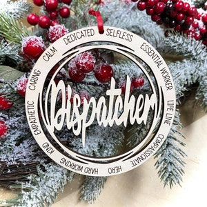 May include: A wooden Christmas ornament with the word "Dispatcher" in a decorative font. The ornament is surrounded by a circle of words describing the qualities of a dispatcher, including "calm", "dedicated", "selfless", "essential worker", "life line", "compassionate", "hero", and "hard working".