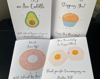 May include: Four white greeting cards with cute food illustrations. The cards feature an avocado, muffin, donut, and two eggs. Text includes "Sending you an Avo-Cuddle", "There's Muffin Stopping You!", "Donut Give Up!", and "Your Friendship Is Eggcellent".