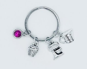 May include: Silver keychain with four charms: a pink rhinestone, a cupcake, a kitchen mixer, and a measuring cup.