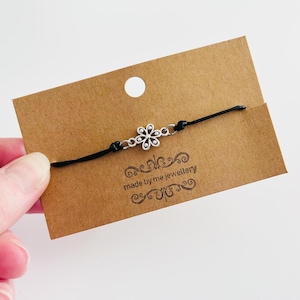 May include: A black string bracelet with a silver flower charm. The bracelet is displayed on a brown card with the text "made by me jewellery".