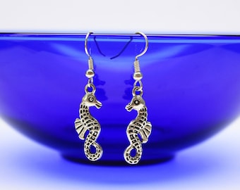 May include: A pair of silver tone dangle earrings featuring a seahorse design. The earrings are hanging from a blue bowl.
