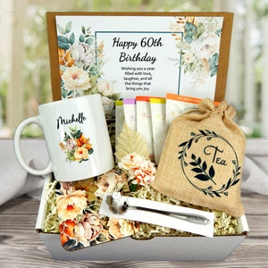 May include: A gift box with a white mug personalized with "Michelle" and floral design. The box includes a card that says "Happy 60th Birthday", tea bags, a burlap bag of tea, a spoon, and decorative flowers.