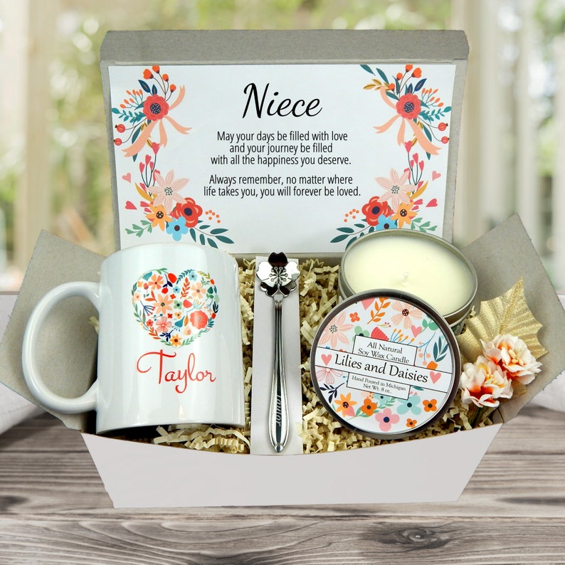 May include: A gift basket containing a white mug with a floral heart design and the name "Taylor", a spoon, a candle, and a card that reads "Niece". The candle is in a tin with a floral design. The basket is filled with shredded paper.