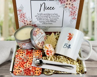 May include: A gift box filled with a white mug with the name "Beth" and a floral design, a candle with a floral design and the text "Lilies & Daisies", a floral coaster, a silver spoon, and a card with the text "Niece" and a floral design.