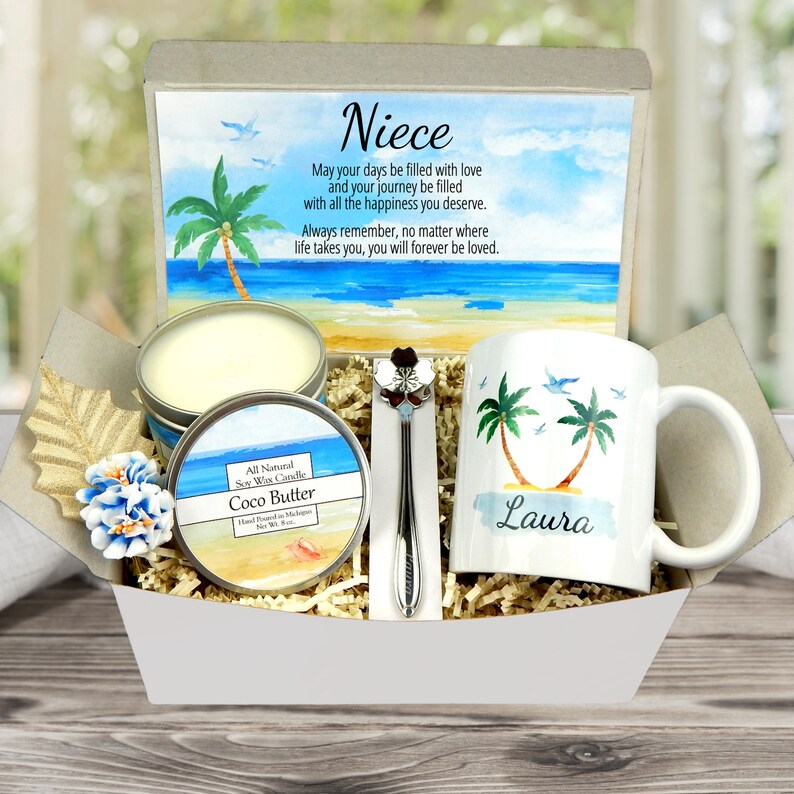 May include: A gift basket containing a white mug with palm trees and the name "Laura", a candle, a tin of Coco Butter soy wax candle, and a decorative spoon. The background features a beach scene with text that reads "Niece".