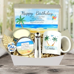 May include: A white gift box filled with a white mug with a tropical beach scene and the name "Laura" on it, a small candle with a tropical beach scene and the text "All Natural Soy Wax Candle Coco Butter", and a silver spoon with a flower design. The box has a card on top with the text "Happy Birthday Wishing you a day filled with joy and love. May this year bring you happiness and peace."