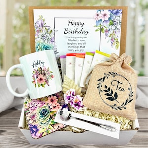 May include: A gift box with a white mug personalized with the name "Ashley" and floral design. The box includes a "Happy Birthday" card, tea bags, a burlap tea bag, a coaster, and a tea spoon. The box is filled with decorative paper.