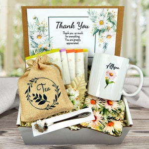 May include: A gift basket with a white mug featuring a daisy and the name "Allison", a burlap tea bag, tea packets, a floral spoon, and a card that says "Thank You". The basket includes a floral coaster and is decorated with daisies.