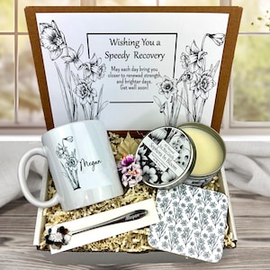 May include: A gift box with a white mug, a candle, a spoon, and a coaster. The mug and spoon are personalized with the name "Megan". The box includes a card with the text "Wishing You a Speedy Recovery". The candle is labeled "Sophora In Bloom".