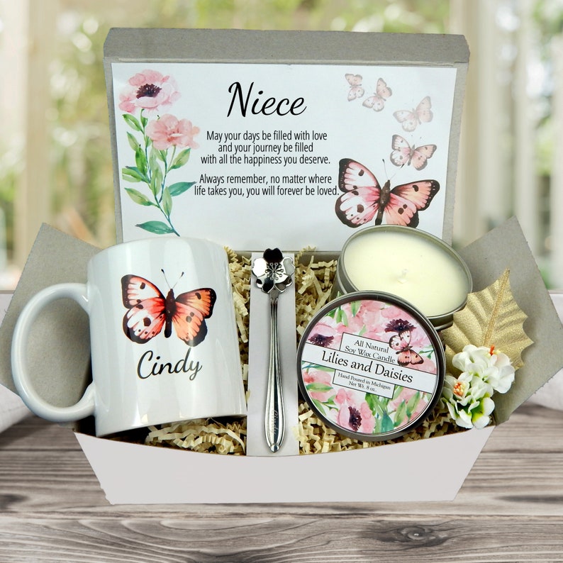 May include: A gift basket with a white mug featuring a butterfly and the name "Cindy". The basket includes a candle, a decorative spoon, and a card with the word "Niece" and a message. The theme is butterflies and flowers.