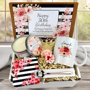 May include: A gift box with a floral theme, containing a mug with "Amanda" and floral design, a candle, a coaster, and a spoon. The box includes a card that says "Happy 30th Birthday". The color scheme includes pink, white, and black.