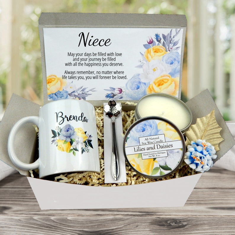 May include: A gift basket containing a white mug with floral design and the name "Brenda", a candle, a spoon, and a card with the word "Niece" and a floral design. The basket includes a "Lilies and Daisies" candle.
